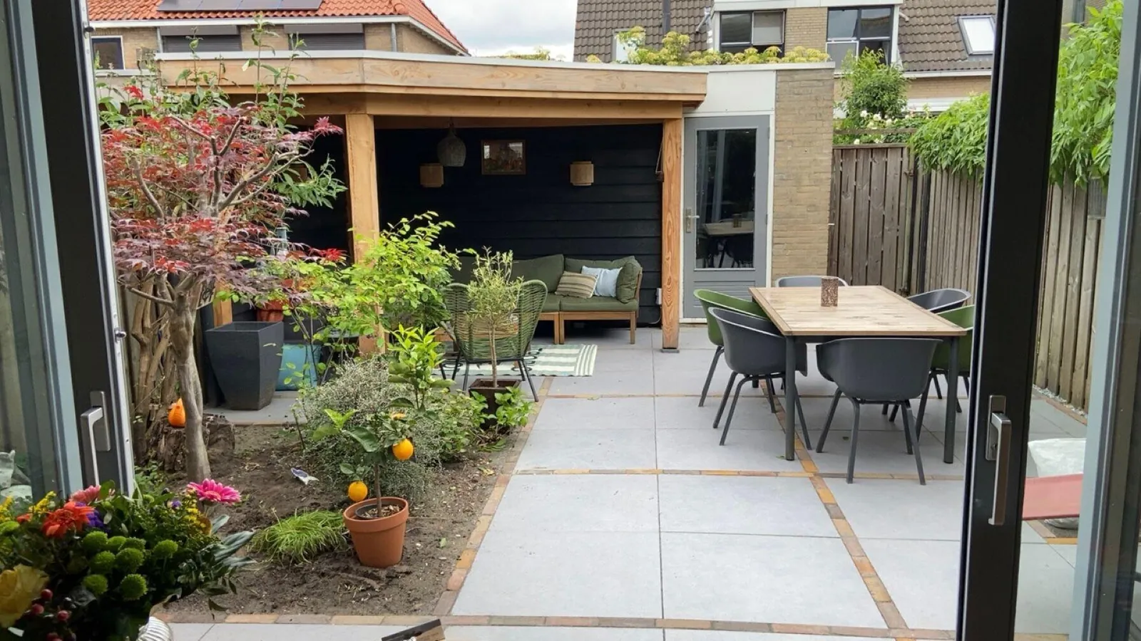 Achtertuin met nieuwe grote grijze tegels, een houten overkapping met loungebank en een tuintafel